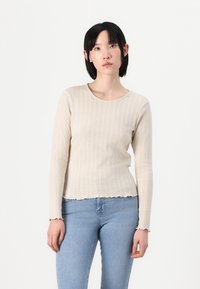 Beige ribbete langermet topp med bølget kant og rund hals, laget av et mykt, teksturert stoff. Parret med lyseblå jeans.