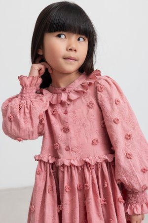 Jeune fille aux longs cheveux noirs, portant une robe rose texturée avec des détails floraux, pose de manière réfléchie sur un fond uni.