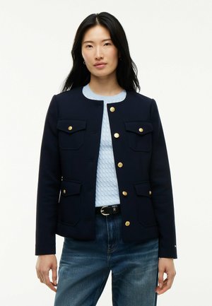 Mujer con cabello largo y negro lleva una chaqueta azul marino con botones dorados sobre un suéter azul claro con textura y unos vaqueros azules.