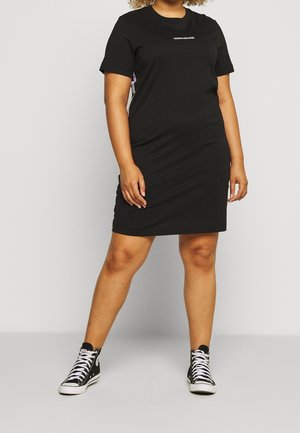 Kobieta stojąca, ubrana w czarną sukienkę Calvin Klein z krótkim rękawem i czarne wysokie tenisówki z białymi sznurowadłami na jednolitym tle.