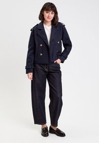 Veste croisée marine avec boutons dorés, larges revers et épaulettes, associée à un pantalon large en denim foncé et des mocassins noirs.