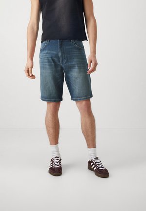 Pantalones cortos de mezclilla en azul medio con un acabado ligeramente desgastado, diseño de cinco bolsillos y puños rematados. Llevados con zapatillas oscuras y calcetines blancos.
