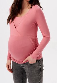 Długi rękaw bluzki ciążowej w miękkim różowym kolorze, z krzyżującym się przodem i marszczonym bokiem dla rozciągliwości, w połączeniu z szarymi dżinsami.