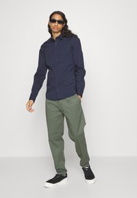 Camicia blu navy con maniche lunghe, abbinata a pantaloni larghi verdi oliva e sneaker nere con suola bianca.