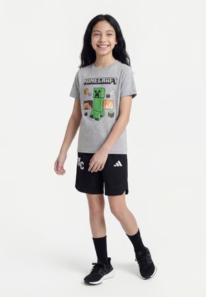 Smilende pige med langt sort hår iført grå Minecraft T-shirt, sorte træningsshorts, sorte sokker og sorte sneakers, stående på hvid baggrund.