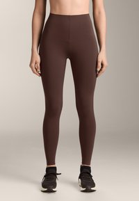 Szűk, magas derekú barna leggings, fekete bokazoknival és fekete sportcipővel viselve, egyszerű háttér előtt.