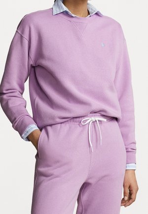 Personne portant un sweat lilas et un pantalon de survêtement assorti avec un cordon blanc, superposés sur une chemise bleue à col, les mains partiellement dans les poches.