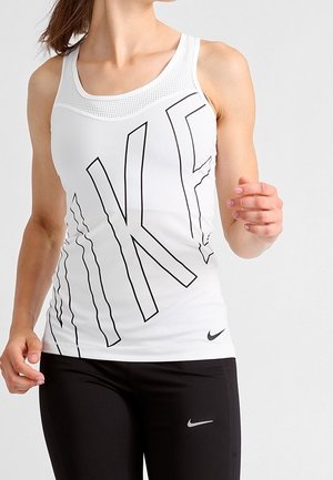 Kobieta ubrana w biały top Nike z dużymi czarnymi literami i czarne legginsy Nike, stojąca na jednolitym szarym tle.