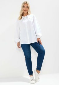 Biała koszula z guzikami, z dużym kołnierzem, bufiastymi rękawami i luźnym krojem. Połączona z ciemnoniebieskimi dżinsami i białymi trampkami.