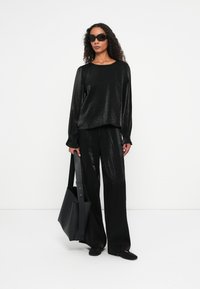 Schwarzes schimmerndes Langarm-Oberteil und weite Hosen mit elastischen Bündchen. Das Modell hält eine große schwarze Tragetasche und trägt dunkle Sonnenbrillen.
