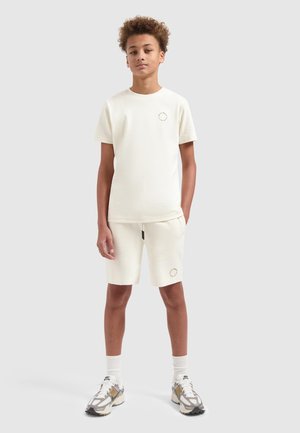 Tienerjongen die staat, gekleed in een crèmekleurig T-shirt met korte mouwen en bijpassende korte broek, met Nike-sneakers en witte sokken, tegen een lichte achtergrond.