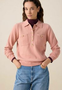 Pull en tricot rose clair avec un col, doté de deux poches avant, de boutons dorés et d'accents côtelés. Porté par-dessus un col roulé sombre.