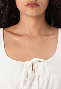 White, lightweight fabric top with a scoop neckline. Drawstring detail at the front and soft texture. Smooth skin tones visible.