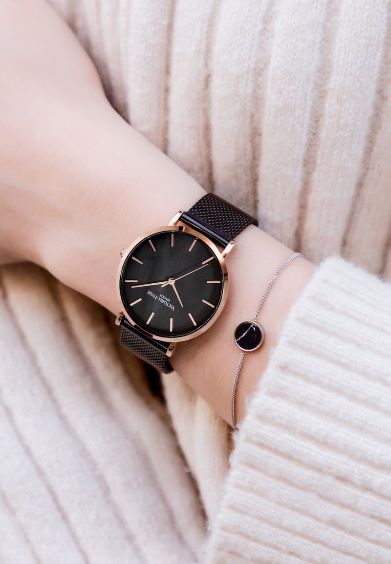Reloj negro con una esfera redonda, acentos dorados y una correa de malla; acompañado de una delicada pulsera de plata con un pequeño detalle circular.
