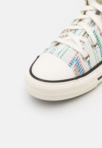 Zapatilla de lona blanca con un patrón de rayas multicolores, que presenta una puntera redondeada, ribete negro y cordones blancos. Suela de goma con detalle texturizado.