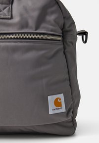 Szürke nylon hátizsák, cipzáras zsebbel, texturált anyaggal, négyzet alakú logóval, narancssárga grafikával és "carhartt" felirattal.