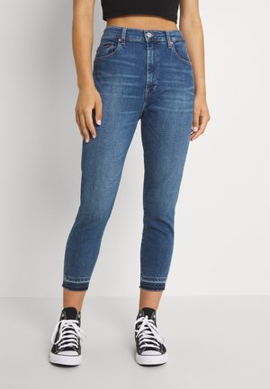 Kvinde iført blå højtaljede cropped skinny jeans og sorte høje sneakers med hvide snørebånd, stående mod en ensfarvet lys baggrund.