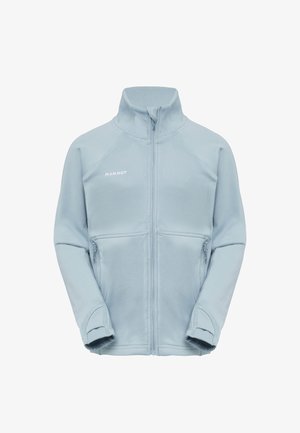 Jeune fille portant une veste zippée bleu clair et un short bleu, souriante avec les mains détendues le long du corps sur un fond uni.