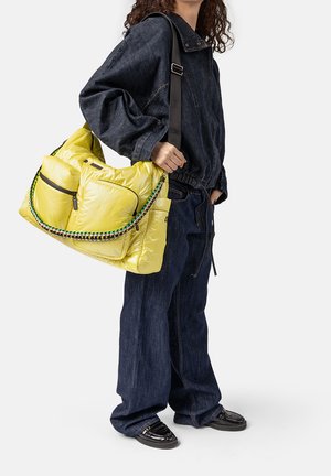 Persoon in een donker denim outfit die een grote gele, puffy schoudertas vasthoudt met voorvakken en een meerkleurige gevlochten riem.