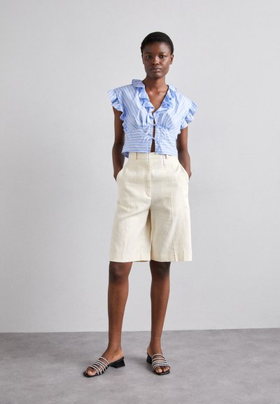 Blouse rayée bleu clair avec des manches volantes, design cropped, associée à un short beige jusqu'aux genoux et des sandales noires avec des accents métalliques.