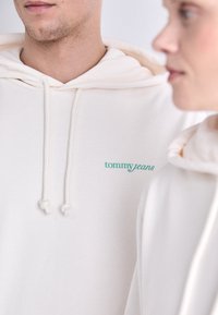 Des sweats à capuche couleur crème en tissu doux, avec un logo turquoise "tommy jeans" et des accents de cordon. Design simple, ajusté.