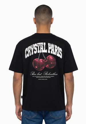 Persona che indossa una maglietta nera con grande testo bianco "CRYSTAL PARIS" e due ciliegie rosse nella grafica sulla schiena.