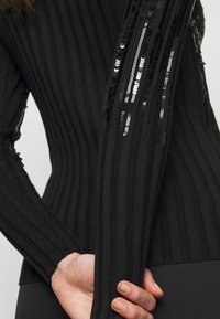 Suéter negro de punto acanalado con mangas largas, que presenta lentejuelas negras a lo largo de las mangas, tela texturizada y silueta ajustada.