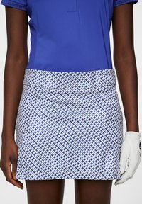 Blåt figursydd piké med korta ärmar, kombinerad med en mönstrad vit kjol med blå geometriska mönster. Håller en vit golfhandske.