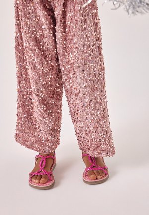 Pantalon recouvert de sequins roses associé à des sandales à brides tressées rose vif sur un fond clair.