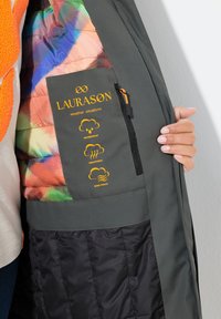 Chaqueta tipo puffer con un forro interior colorido, tela exterior gris, bolsillo con cremallera e íconos climáticos que indican características impermeables, transpirables y a prueba de viento.