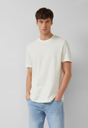 Jonge man met kort bruin haar, draagt een eenvoudig wit T-shirt met korte mouwen en lichtblauwe spijkerbroek, staand met handen in de zakken.