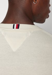 Close-up van een persoon die een witte gebreide trui draagt met een rood-wit-blauwe streepdetail bij de achterkant van de halslijn en een zilveren ringoorbel.