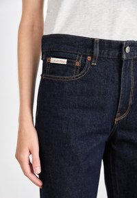 Mörka denimjeans med orange sömmar, som har en framficka med en röd och vit Calvin Klein-lapp samt en metallknappsstängning.