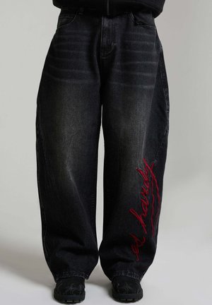 Weit geschnittene schwarze Jeans mit rotem, vertikal auf dem rechten Bein gesticktem Text, kombiniert mit schwarzen Schuhen und dunklem Oberteil.