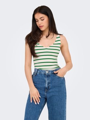 Ung kvinde med langt mørkt hår iført en hvid tanktop med grønne vandrette striber og blå højttaljede jeans, den ene hånd i lommen.