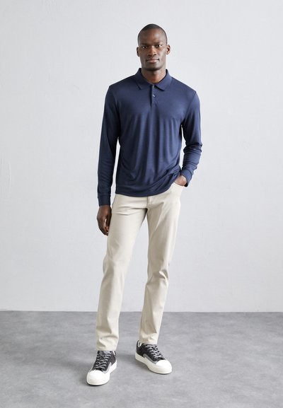 Marineblaue Langarm-Polohemd, weicher Stoff, Drei-Knopf-Leiste, kombiniert mit beigen Slim-Fit-Hosen und grauen Turnschuhen mit weißen Sohlen.