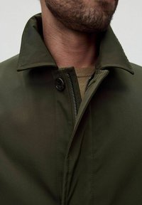 Vihreä takki pehmeällä pinnalla, jossa on leveä kaulus, napitettu sulkeminen ja vetoketjuelementti. Näkyvä sisäkerros neutraalissa värissä.