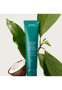Aveda Botanical Repair bond-building styling crème i en turkos 150 ml tub, omgiven av gröna blad och en halvmandel, med en mjuk konsistens.
