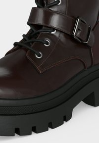 Gros plan d'une botte en cuir brun foncé avec des lacets noirs, une lanière en cuir et une boucle argentée, ainsi qu'une semelle noire épaisse texturée avec des rainures profondes.