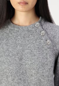 Grauer Pullover aus Strickstoff, mit Rundhalsausschnitt und drei dekorativen Knöpfen mit floralem Design an einer Schulter.