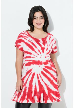 Uśmiechnięta kobieta o ciemnych włosach, nosząca duże kolczyki-koła i krótką sukienkę z czerwono-białym wzorem tie-dye z falbaną na dole.