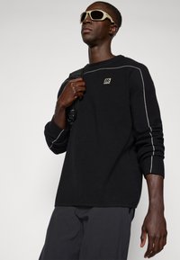 Maglione nero con cuciture bianche, caratterizzato da un logo quadrato sul petto. Il modello tiene uno zaino nero texture, indossando occhiali da sole beige.