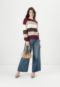 Pull rayé bordeaux, crème et marron, associé à un pantalon en denim large, tenant un sac beige à poignée tressée.
