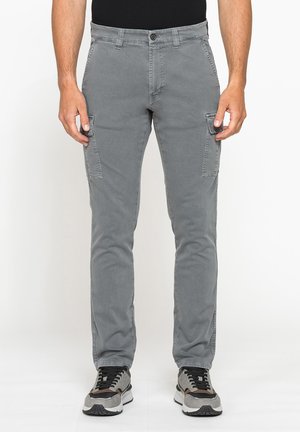 Uomo che indossa pantaloni cargo grigi slim fit e scarpe da ginnastica nere e grigie, in piedi contro uno sfondo bianco uniforme.