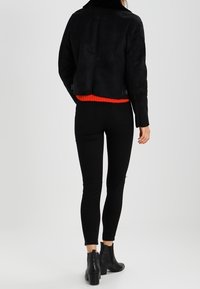 Czarna zamszowa krótka kurtka z wysokim kołnierzem i czerwonym dzianinowym pasem, połączona z czarnymi obcisłymi dżinsami i botkami za kostkę.