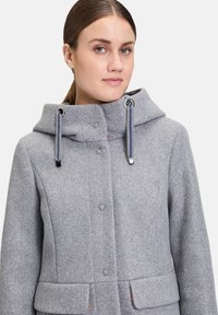 Grauer Wollmantel mit hohem Kragen, Druckknöpfen und zwei Vordertaschen. Mit abnehmbarer Kapuze mit Kordelzügen und metallischen Akzenten.