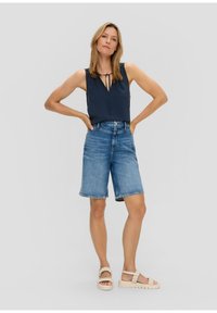 Top senza maniche blu navy con dettaglio a fiocco, abbinato a bermuda in denim azzurro chiaro. Indossato con sandali intrecciati color crema. Sfondo neutro.