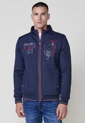 Chaqueta de navy con cremallera y cuello alto, con acentos rojos y blancos, gráficos de texto y dos bolsillos laterales. Hecha de una mezcla de algodón.