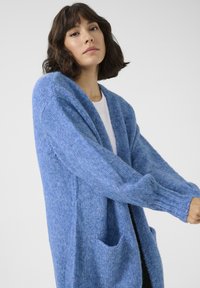 Cárdigan de punto azul con un acabado texturizado, que presenta un escote en V, mangas largas y dos bolsillos frontales. Material suave y corte relajado.