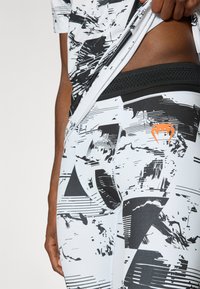 Λευκό αθλητικό μπλουζάκι με μαύρα αφηρημένα σχέδια, φορεμένο πάνω από μαύρο κολάν συμπίεσης. Διαθέτει πορτοκαλί λογότυπο στο πλάι του κολάν.